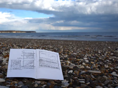 An open sketchbook lies on a pebble beach with thumbnail sketches on its pages